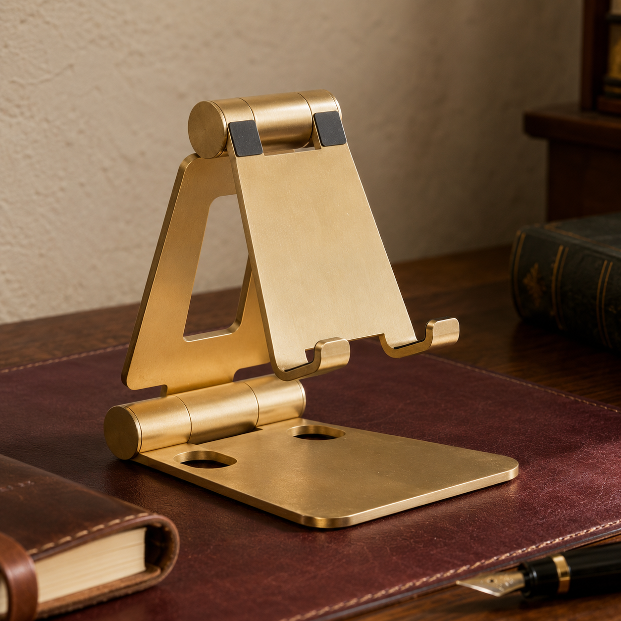 Brass-finish folding cell phone stand holding a phone at eye level on a dark wood writing desk
