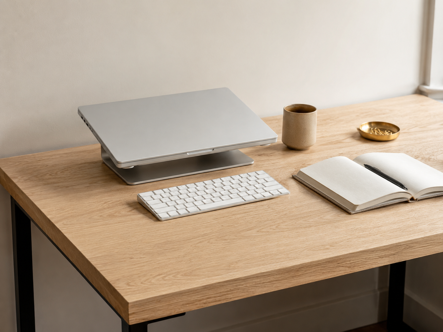A minimalist laptop-only desk setup with a closed silver MacBook on a clean white oak desk, with a wireless mechanical keyboard, ceramic mug, and linen notebook