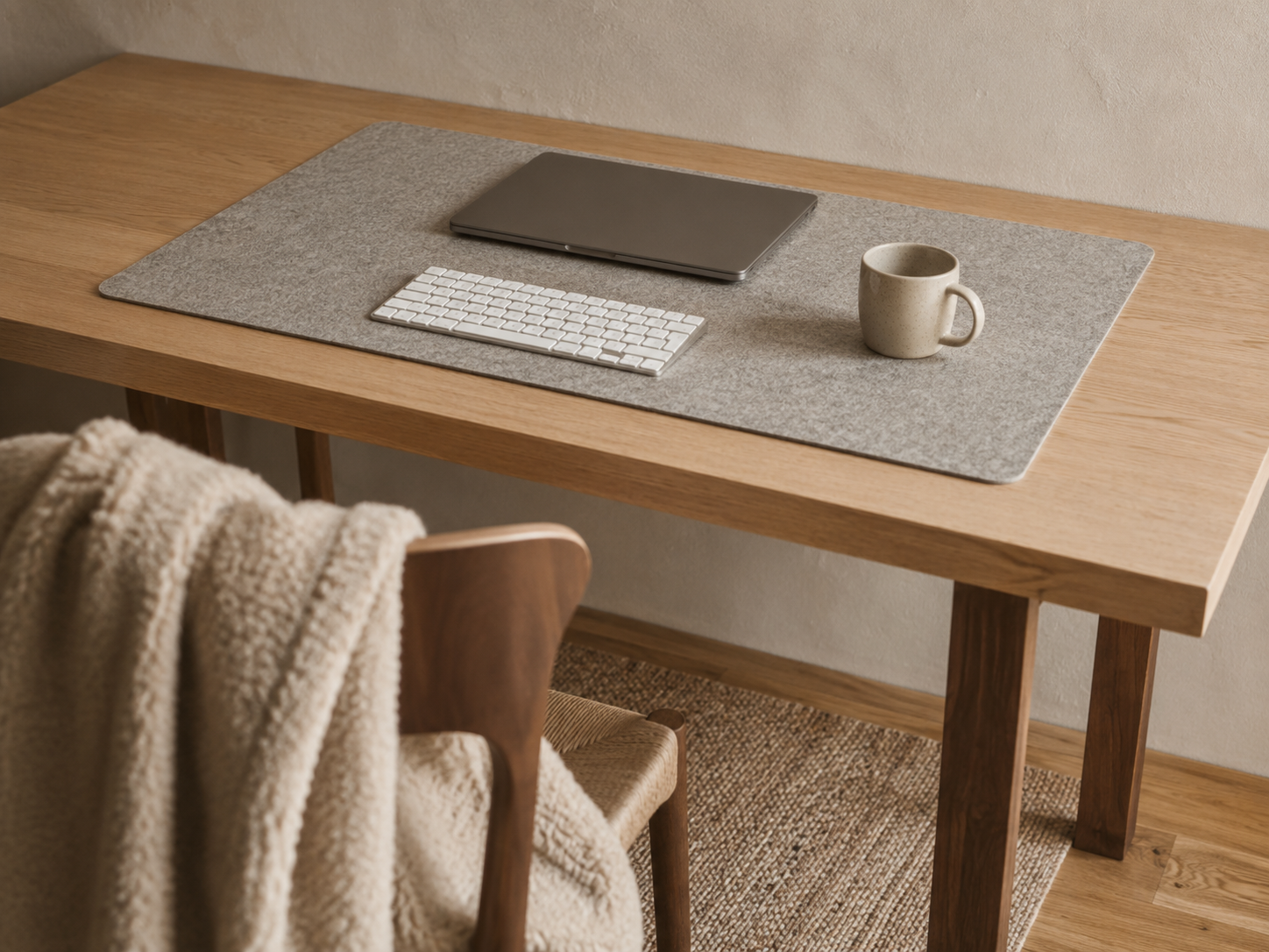 A cozy desk setup with a wool felt deskmat in warm gray, a slim mechanical keyboard with cream keycaps, and a layered jute rug visible beneath the chair on hardwood flooring